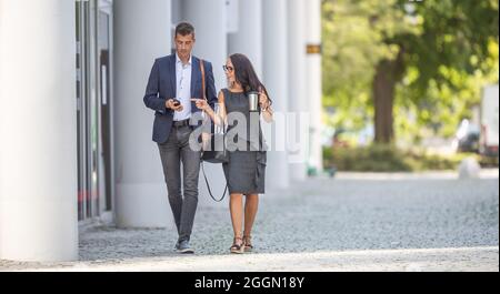 Two office workers walk to work discussing news on the mobile phone. Stock Photo