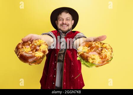 Happy smiling man dressed in traditional Bavarian costume with yummy pretzels isolated over yellow background. National cuisine, holidays, traditions Stock Photo