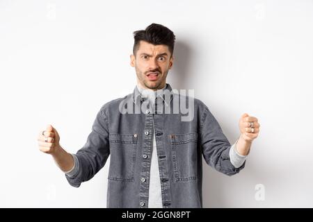 Skeptical young man showing big size object and frowning doubtful, standing reluctant on white background Stock Photo