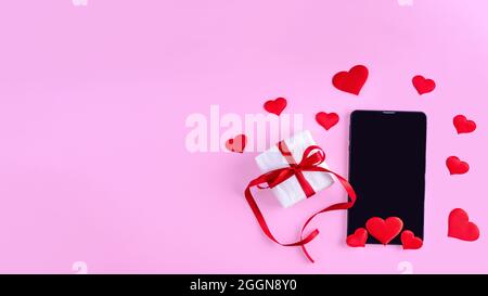 Online congratulations concept. Black blank tablet or phone screen with red hearts shape and gift in white wrapping paper with red ribbon on pink back Stock Photo