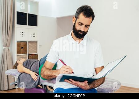 Portrait of a bearded doctor taking notes on his clipboard of the history of his patient who is lying on the stretcher behind him. Health concept Stock Photo