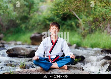 Portrait of a Japanese taekwondo boy sitting in nature and doing meditation exercises. The young asian black belt karate sitter. Stock Photo