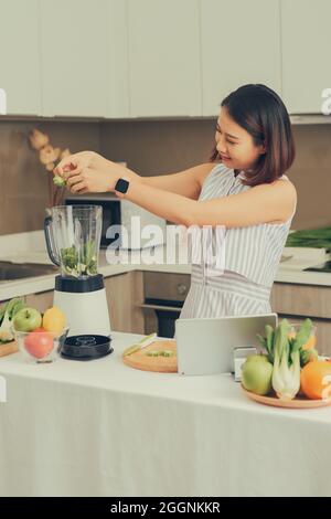 Smiling vegan Asian woman making a smoothie with fruit and vegetable in the kitchen