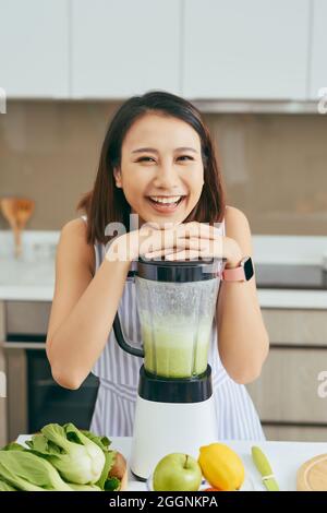 Smiling vegan Asian woman making a smoothie with fruit and vegetable in the kitchen Stock Photo