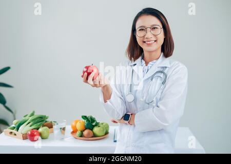 Smiling nutritionist in her office, she is holding a green apple and showing healthy vegetables and fruits, healthcare and diet concept Stock Photo