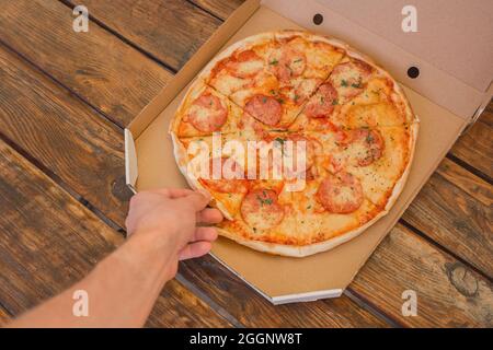 The guy's hand takes a pizza in cardboard box of against the background of a wooden table. Delicious fast food. Stock Photo