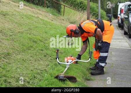 Workman in full protective clothing adjusting a petrol strimmer ready to cut a sloping grass verge
