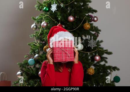 A funny child in a Santa Claus hat hides his face behind a box with a Christmas present. Little girl on the background of a Christmas tree. Stock Photo