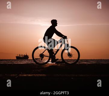 Silhouette of man riding bicycle along the coastal road during the sunset Stock Photo