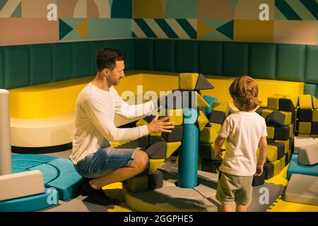 Dad and son are building castle of soft colorful shapes in children's center. Educational classes and games. Recreation and entertainment. Game and de Stock Photo