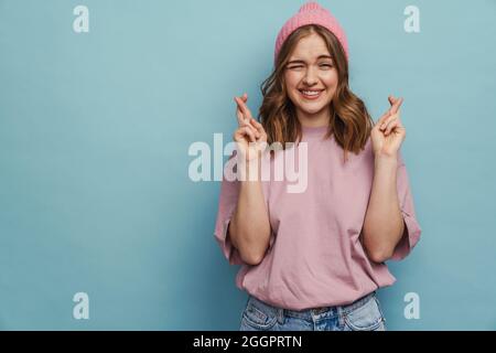 Young happy woman squeezing her eyes and keeping fingers crossed isolated over blue background Stock Photo