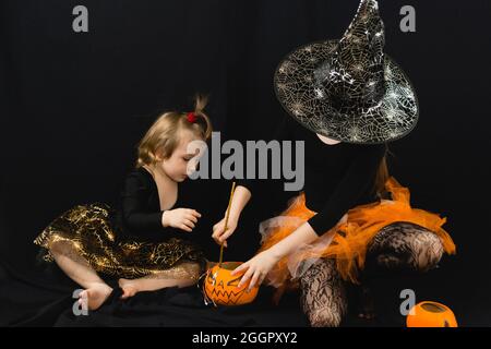 Two sisters in witch costumes, cooking a magic potion in a bucket-pumpkin. A black blouse, a hat with cobwebs, an orange skirt. Black background Stock Photo