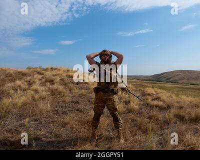 Photo of a mercenary sniper in camouflage clothes under the scorching sun. He stands with a rifle and surveys the area. Concept of a professional army Stock Photo