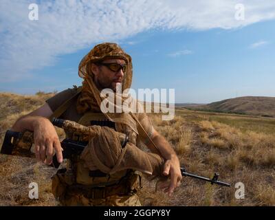 A mercenary sniper in camouflage clothes under the scorching sun. He stands with a rifle and surveys the area. Concept of a professional army during a Stock Photo