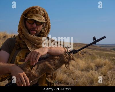 Close-up photo of a mercenary sniper in camouflage clothes under the scorching sun. He stands with a rifle and looking to camera. Concept of a profess Stock Photo