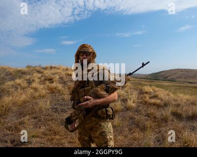 Sniper-mercenary in camouflage clothes under the scorching sun. He stands with a rifle and surveys the area. Concept of a professional army during arm Stock Photo