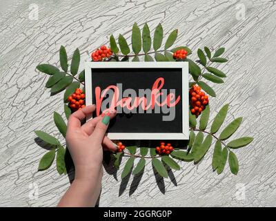 Text Danke, thank you in German language. Orange rowan berry with green leaves around blackboard, chalk board. Hand holds paper word, text Thank You Stock Photo