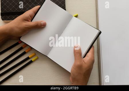 hands of a person holding an open notebook with blank sheets and colored pencils in the background on a desk, office instruments in workspace, detail Stock Photo