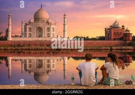 Taj Mahal Agra at sunset with moody sky and water reflection enjoyed by tourist couple sitting at Mehtab Bagh beside river Yamuna Stock Photo