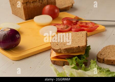 closeup of delicious sandwich ready to eat with cheese, lettuce, fresh tomato, on a table, in the background a chopping board with fresh cut vegetable Stock Photo