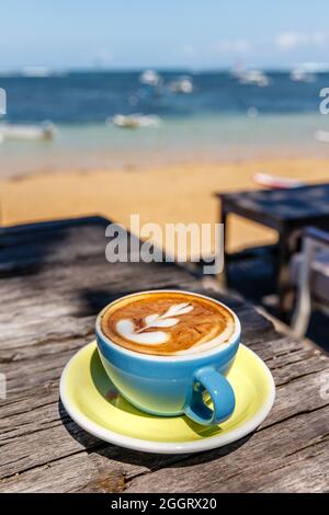 Cup of cappuccino standing on a wooden table in a cafe, view of the sea and a beach in the background. With space. Stock Photo