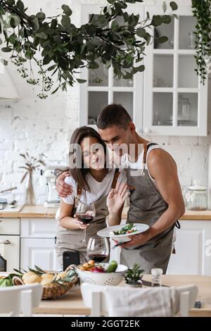 Happy millennial caucasian couple making video call with smartphone while preparing romantic dinner in the kitchen. Cheerful young people smiling at t Stock Photo