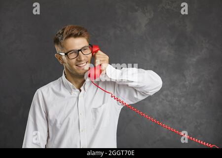 Studio portrait of a happy young man talking on red landline telephone on gray background. Stock Photo