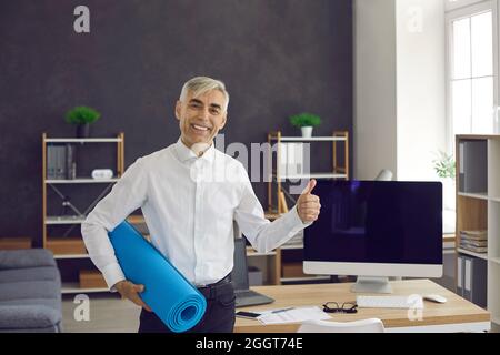 Happy fit senior man standing in office, holding yoga mat, giving thumbs up and smiling Stock Photo