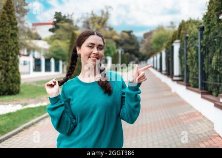 Portrait of happy female student with pigtails stands on the street and pointing at the right side. Outdoor, fence. The concept of admission of studen Stock Photo