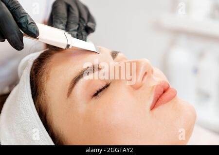 Procedure at the beauty salon. Cosmetologist cleans the client's forehead using a ultrasonic device. Close up. Concept of professional cosmetology. Stock Photo