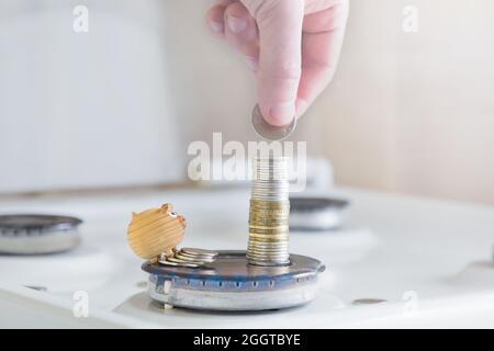 the concept of economy and investment .a man's hand puts a coin on a stack of money on the background of a gas burner and a small toy pig. Stock Photo