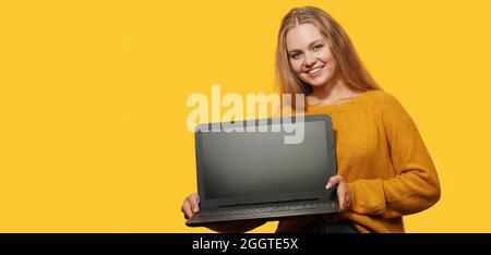 Blonde Woman Holding Laptop With Blank Screen over yellow background. Copy space.    Female holds laptop screen forward with mock up. Stock Photo