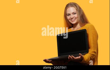 Blonde Woman Holding Laptop With Blank Screen over yellow background. Copy space.    Female holds laptop screen forward with mock up. Stock Photo