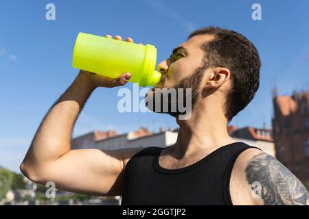 Sports man drinks water or protein cocktail outdoors. Handsome muscular guy with tattoo goes in for sports Stock Photo