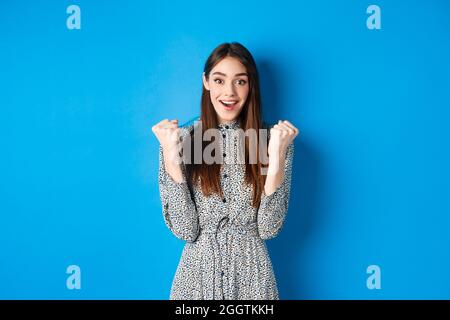 Excited woman looking relieved and hopeful, making fist pump and smiling, winning prize, celebrating achievement or victory, standing on blue Stock Photo
