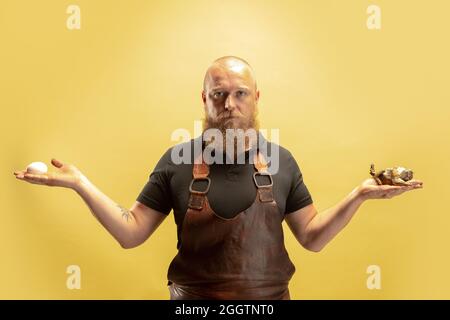 Comic portrait of muscular bearded bald man, blacksmith in leather apron or uniform isolated on yellow studio background. Concept of labor, retro Stock Photo