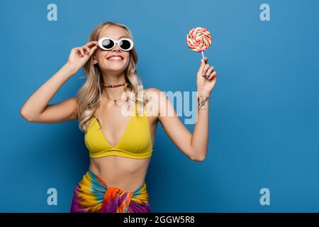 smiling young woman in yellow bikini top and sunglasses holding sweet lollipop on blue Stock Photo