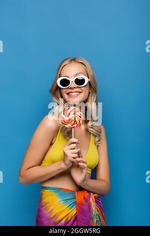 smiling young woman in yellow bikini top and sunglasses holding sweet lollipop isolated on blue Stock Photo