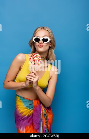 young woman in yellow bikini top and sunglasses pouting lips while holding sweet lollipop isolated on blue Stock Photo