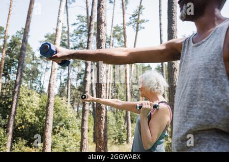 Side view of elderly woman training with dumbbells near blurred african american husband in forest Stock Photo