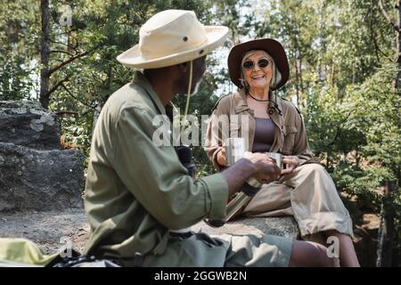 Smiling traveler holding cups near blurred african american husband with thermos in forest Stock Photo