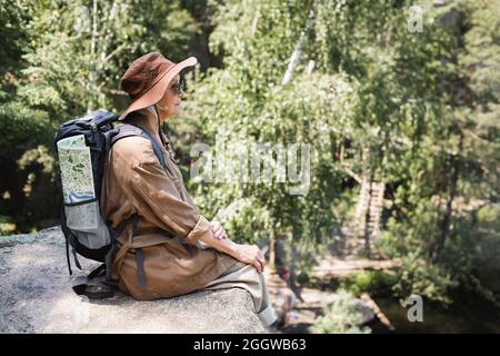 Side view of elderly woman with backpack and map sitting on cliff Stock Photo