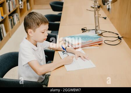 The boy sits in the library and writes in a notebook at the table. Preparing for homework. a good student. Schoolboy loves to study. Stock Photo