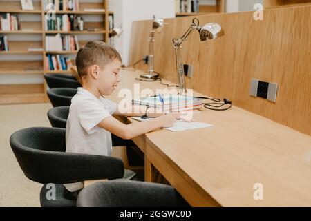 The boy sits in the library and writes in a notebook at the table. Preparing for homework. a good student. Schoolboy loves to study. Stock Photo