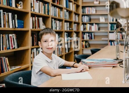 The boy sits in the library and writes in a notebook at the table. Preparing for homework. a good student. Schoolboy loves to study. Free space at sch Stock Photo