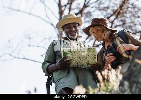 Low angle view of cheerful interracial couple of hikers looking at map in forest Stock Photo
