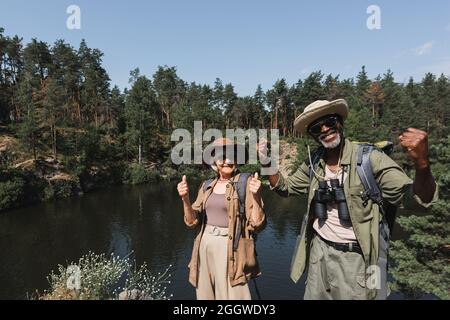Positive multiethnic couple with backpacks gesturing near lake and forest Stock Photo
