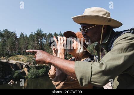 Side view of african american hiker pointing with finger near smiling wife with binoculars outdoors Stock Photo