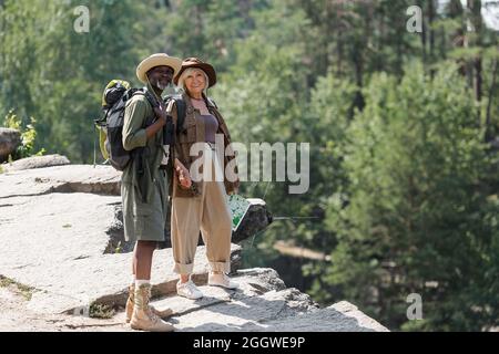 Cheerful multiethnic senior travelers with backpacks standing on rock Stock Photo