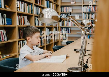 Schoolboy sitting on table and reading book in library at school. Preparing for homework. a good student. The boy loves to read. Free space at the sch Stock Photo